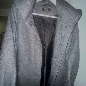 Grå kappa från ONLY med huva XS - Säljer en snygg grå kappa från ONLY i storlek XS. Jackan har en stor huva, knappar framtill och är helfodrad. Materialet är mjukt och känns ulligt, perfekt för kyliga dagar. Har en ficka. Defekt bild 3: sönder precis vid fickan, men hindrar inte funktionen.