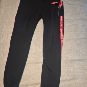 Alpha Industries svarta mjukisbyxor L - Svarta mjukisbyxor från Alpha Industries med röda detaljer längs sidorna där det står 'REMOVE BEFORE FLIGHT'. Byxorna har resår i midjan och mudd vid benslut. Tillverkade i mjukt material, perfekt för chill eller streetwear. Snygg detalj med röd nyckelring vid fickan.