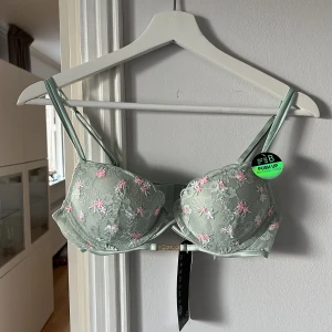 Grön korsett-bh med blommig spets - Snygg korsett-bh (push-up) från New Yorker i ljusgrön färg med broderade rosa och vita blommor på spetsen. Den är helt ny och aldrig använd. Köpte för 160kr