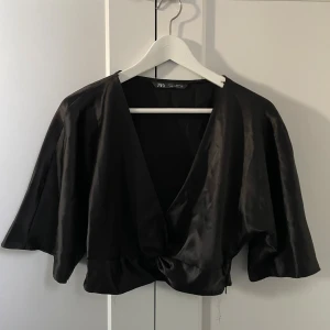 Svart omlott blus från Zara - Snygg svart omlott blus från Zara med glansigt material och vida, korta ärmar. Blusen har en djup v-ringning och knytdetalj framtill, den är kort!