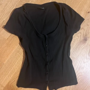 Svart figurnära ribbad topp från Gina Tricot - Svart ribbad topp från Gina Tricot med korta ärmar och knappar hela vägen framtill. Toppen har en djup rundad ringning och är figurnära i modellen. Perfekt att styla med jeans eller kjol för en enkel och snygg look.