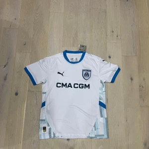 Vit Marseille fotbollströja Puma - Snygg vit Olympique de Marseille matchtröja från Puma med blå detaljer vid krage och ärmslut. Tryckt klubbmärke och sponsorlogga på bröstet. Tillverkad i lätt och ventilerande funktionsmaterial, perfekt för träning eller match.
