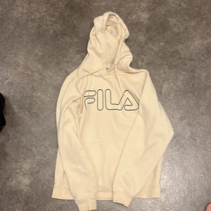 Beige hoodie från FILA med logga - Snygg beige hoodie från FILA med stor svart logga framtill. Tröjan har huva med snörning och en klassisk känguruficka. Perfekt för chill dagar eller när du vill ha en enkel och stilren look.
