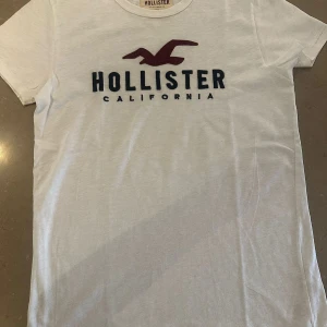 Vit t-shirt från Hollister S - Vit t-shirt från Hollister med klassisk logga och texten 'Hollister California' framtill. T-shirten har rund hals, korta ärmar och är gjord i mjuk bomull. Perfekt basic med snyggt tryck och relaxed vibe.
