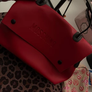 Röd handväska från Moschino i skinn - Säljer en snygg röd handväska från Moschino i slätt skinn med präglad logga framtill. Väskan har svarta detaljer, praktiskt handtag och rymligt fack med innerficka. Perfekt för dig som vill sticka ut med en statementväska.