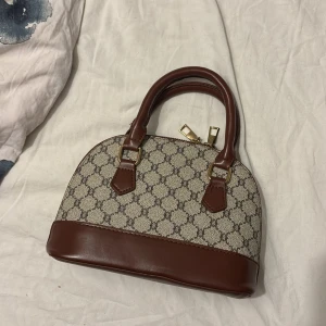 Brun och beige handväska med mönster - Snygg handväska i beige canvas med klassiskt monogrammönster och bruna skinn-detaljer. Väskan har guldfärgade metalldetaljer, två handtag och avtagbar axelrem. Stängs med dragkedja och har rymligt ljust innerfack.