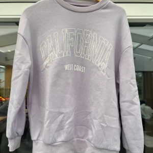 Ljuslila sweatshirt med California-tryck - Säljer en ljuslila sweatshirt med trycket 'California West Coast' framtill. Tröjan har rund halsringning, långa ärmar och ribbade muddar vid ärmslut och nederkant. Perfekt för en avslappnad och trendig look.