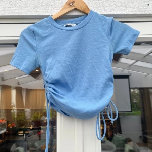 Blå ribbad topp med snörning - Snygg ljusblå ribbad topp från Bik Bok med korta ärmar och rund hals. Toppen har trendig snörning i sidan som gör att du kan justera passformen och skapa en cool rynkad effekt. Perfekt att styla med jeans eller kjol för en fräsch look.