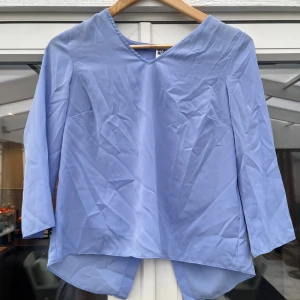 Ljusblå blus med omlott-detalj - Säljer en ljusblå blus från Vero Moda med v-ringning och trekvartsärm. Blusen har en snygg omlott-detalj baktill och är längre bak än fram för en cool look. Perfekt att styla med jeans eller kjol.