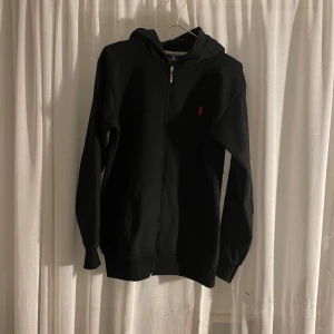 Svart hoodie från Polo Ralph Lauren - Säljer en stilren svart hoodie från Polo Ralph Lauren. Den har en dragkedja framtill och en liten röd logga på bröstet.  Nopprig, annars inga hål eller liknande. Saknar dock band till luvan.