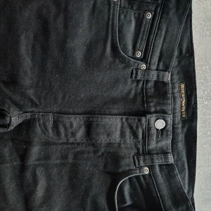 Svarta jeans från Nudie Jeans 29/32 - Snygga svarta jeans från Nudie Jeans i klassisk femficksmodell. Jeansen har en stilren look med svarta sömmar och metallknappar. Tillverkade i mjukt denimtyg som sitter skönt och passar till det mesta. Perfekta för dig som gillar en enkel och clean stil. Nästan aldrig användt