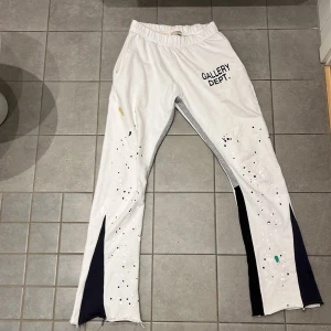 Gallery Dept mjukisar -  vita byxor från Gallery Dept. med svart text på framsidan, färgstänk i svart, vitt, grönt och gult samt svarta paneler längs benen. Byxorna har elastisk midja med snörning och är tillverkade i 100% bomull. Råa kanter nertill för en edgy look.