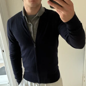 Mörkblå kofta från Riley med dragkedja - Snygg mörkblå kofta från Riley i storlek S. Koftan har en stilren design med hel dragkedja framtill, ribbade muddar och rund halsringning. Perfekt för lager-på-lager och passar till både skjorta och t-shirt. Bra skick, passar även XS