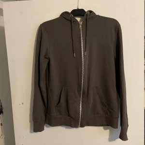 Mörkbrun hoodie från The Black Label - Mörkbrun hoodie med dragkedja från The Black Label Collection. Tröjan har huva med snörning, två fickor framtill och är tillverkad i mjuk bomull. Perfekt för chill dagar eller när du vill ha en enkel och stilren look.