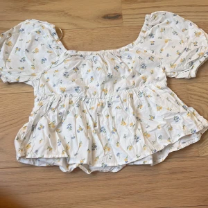 Vit blommig blus med puffärm - Supersöt vit blus med små gula och blå blommor, puffiga korta ärmar och volang nedtill. Blusen har en rundad halsringning med resår och är gjord i mjuk bomull. Perfekt för en somrig look!