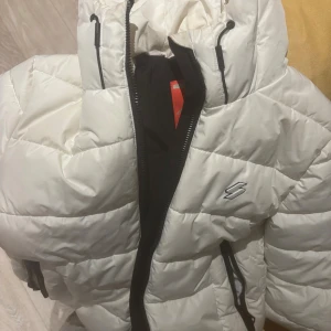 Vit pufferjacka från Superdry - Snygg vit pufferjacka från Superdry Sports Puffer Series. Jackan har svart dragkedja, två fickor med dragkedja och justerbar huva. Perfekt för kalla dagar med sin vadderade design och sportiga look. Logga på bröstet och coola detaljer.   det finns en liten sminkfläck