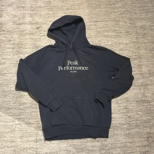 Mörkblå hoodie från Peak Performance - Snygg mörkblå hoodie från Peak Performance med broderad logga på bröstet. Klassisk känguruficka, huva med snörning och mjukt fleecefoder på insidan. Perfekt för kyliga dagar och en avslappnad stil.