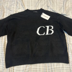 Svart stickad tröja Cole Buxton - Svart stickad tröja från Cole Buxton med vita CB-bokstäver på bröstet. Tröjan har rund halsringning, ribbade muddar och är långärmad. Perfekt för dig som gillar stilrena och cleana plagg med en modern touch.