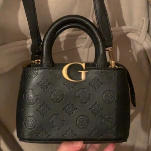 Svart Guess handväska i skinn - Snygg svart handväska från Guess i skinn med präglat logomönster och guldfärgade detaljer. Väskan har både handtag och axelrem samt dragkedja upptill. Perfekt storlek för det viktigaste och en riktigt trendig accessoar. ((Användbar men köpt för 850)).År 2022 på rea så priset kan. diskuteras.