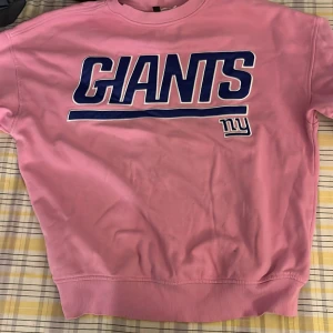 Rosa Giants sweatshirt från H&M Divided - Rosa sweatshirt från H&M Divided med stort blått GIANTS-tryck och NFL-logga framtill. Tröjan har rund hals, ribbade muddar och är i mjuk bomullsmix. Perfekt för dig som gillar sportig stil och vill sticka ut med färg. inklusiv  frakt. stor i storleken 