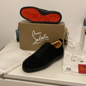 Christian Louboutin svarta sneakers med nitar - Svarta sneakers från Christian Louboutin i mocka med ikoniska röda sulor och svarta nitar på tån. Skorna har snörning och rund tå, samt lyxig läderinsida i beige. Perfekta för dig som vill sticka ut med exklusiv streetstyle.