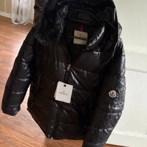 Svart Moncler Maya - Svart pufferjacka från Moncler med glansig finish, huva och dragkedja framtill. Jackan har Moncler-logga på ärmen och innerfoder med serietidningsmönster. Perfekt för kalla dagar och riktigt snygg streetstil.