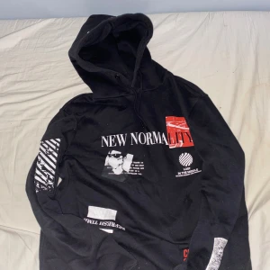 Svart hoodie med tryck New Normality - Svart hoodie med coola tryck i vitt och rött både fram och bak. Texten 'NEW NORMALITY' och grafiska detaljer ger en edgy vibe. Huva med snörning och lång ärm. Perfekt för dig som gillar streetwear och statement-plagg.