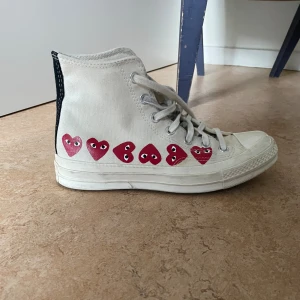 Converse x CDG Play vita sneakers - Säljer ett par vita Converse x Comme des Garçons Play high-top sneakers med röda hjärtan med ögon längs sidan. Skorna har klassisk snörning, rund tå och är tillverkade i canvas. 