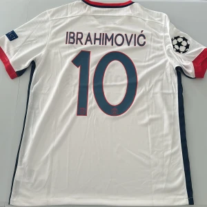 PSG Ibrahimović 10 Champions League tröja - Paris Saint-Germain bortatröja med Ibrahimović 10-tryck, Champions League-märke på ärmen och Fly Emirates sponsor. Vit med röda och marinblå detaljer, tillverkad i lätt Dri-FIT polyester. Klassisk Nike-logga och PSG-emblem på bröstet. Replika, endast handtvätt gäller!