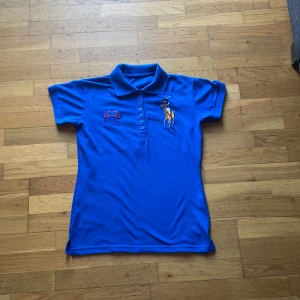 Blå Polo Ralph Lauren t-shirt - Snygg blå t-shirt från Polo Ralph Lauren med klassisk krage och korta ärmar. Framsidan har broderad polospelare och logga, medan baksidan har en stor röd siffra 3. Tillverkad i mjuk bomull, perfekt för en sportig och chill stil. tröjan är lite noppig