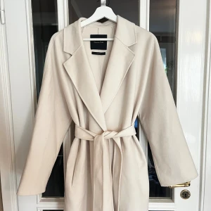 Massimo Dutti beige kappa med knytband - Lyxig beige/krämvit Massimo Dutti kappa med knytband. Det är en ullblandning och kappan är väldigt mjuk och skön. Sån fräsch färg men kommer inte till användning, perfekt som höstjacka. Har små märken på kragen och en liten prick på armen men inget som märks tydligt och bör gå bort i tvätten!