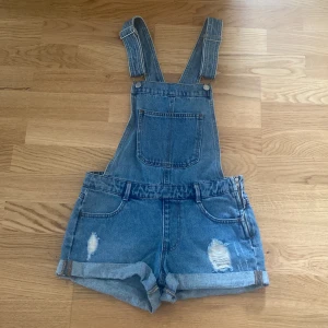Blå jeansshorts hängselshorts - Snygga blå jeansshorts med hängslen och slitna detaljer. Klassisk femficksmodell med uppvikta benslut och stor bröstficka framtill. Perfekt för en chill sommarlook. Justerbara axelband och dragkedja i sidan för enkel påtagning.