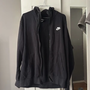 Svart Nike hoodie med dragkedja - Svart hoodie från Nike med vit logga på bröstet. Tröjan har huva med snörning, hel dragkedja och två stora fickor framtill. Insidan är mjuk och fleecefodrad för extra komfort. Perfekt för chill dagar eller när du vill ha en sportig look.