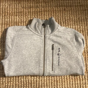 Ljusgrå ziptröja från Sail Racing - Ljusgrå tröja från Sail Racing med hel dragkedja framtill och en bröstficka med dragkedja. Tröjan har hög krage och broderad logga på bröstet. Perfekt för lager-på-lager och chill dagar.