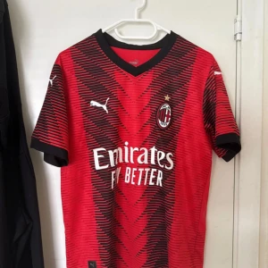 AC Milan matchtröja Puma röd/svart - AC Milan fotbollströja från Puma i rött och svart med grafiskt mönster och klubbmärke på bröstet. Kortärmad modell med svart krage och ärmslut. Tillverkad i lätt och ventilerande funktionsmaterial, perfekt för match eller träning.