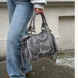 Grå city väska med nitar - Snygg grå axelväska i mocka med mässingsfärgade nitar och dragkedja framtill. Väskan har både handtag och justerbar axelrem