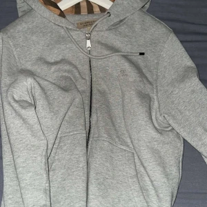Grå hoodie från Burberry med dragkedja - Grå hoodie från Burberry med klassiskt rutigt foder i huvan, dragkedja framtill och snörning vid huvan. Tröjan har broderad logga på bröstet och är tillverkad i mjuk bomull. Perfekt för dig som gillar stilrena och exklusiva plagg.
