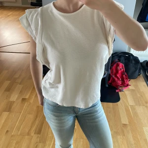 Vit volangblus från Holly & Whyte - Säljer en söt vit blus från Holly & Whyte med volangdetaljer på axlarna. Blusen är ärmlös och har en lätt strukturerad yta. Perfekt till jeans eller kjol för en fräsch look. Använd ett fåtal gånger.