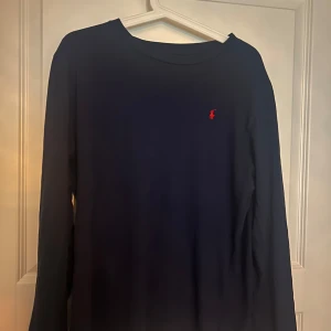 Mörkblå långärmad tröja från Polo Ralph Lauren - Säljer en mörkblå långärmad tröja från Polo Ralph Lauren med den klassiska röda loggan broderad på bröstet. Tröjan har en enkel och stilren design med rund halsringning och är tillverkad i mjuk bomull.