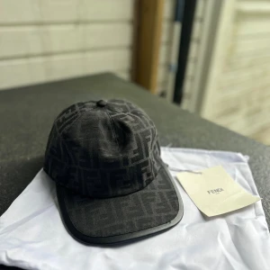 Svart Fendi keps med logomönster - Snygg svart keps från Fendi med det klassiska FF-logomönstret över hela kepsen. Kepsen har böjd skärm med svart kant och justerbar rem baktill med Fendi-etikett. Materialet är mjukt och känns lyxigt, perfekt för dig som gillar designeraccessoarer.