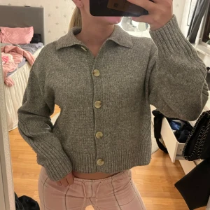 Grå stickad kofta med knappar - Mysig grå stickad kofta med stora knappar framtill och breda ribbade muddar. Koftan har en klassisk krage. Enkel att matcha med olika outfits antigen stängd som på bild eller öppen med nått fint linne under.