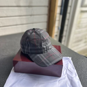 Rutig Burberry keps i grått och rött - Snygg keps från Burberry med klassiskt rutigt mönster i mörkgrå och röda toner. Kepsen har böjd skärm och justerbar rem bak. Tillverkad i bomull med broderad Burberry-logga framtill. Perfekt för dig som vill ha en stilren och trendig accessoar.