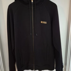Svart hoodie från BOSS med dragkedja - Svart hoodie från BOSS med guldfärgad logga på bröstet. Tröjan har hel dragkedja, huva med snörning och två fickor framtill. Perfekt för en chill och stilren look.