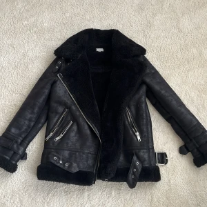 Svart teddyjacka från Warehouse - Snygg svart teddyjacka från Warehouse med bred krage, dragkedjor och spänndetaljer vid ärmslut och midja. Jackan har mjukt foder och är perfekt för kalla dagar. Flera fickor med dragkedja.