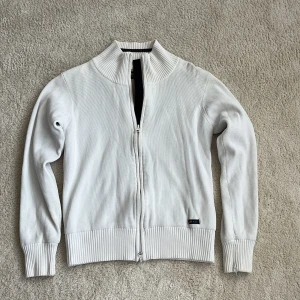 Vit kofta från Newport med dragkedja - Vit ribbstickad kofta från Newport med hög krage och hel dragkedja framtill. Tröjan har långa ärmar, breda muddar och en liten logotyp nertill. 