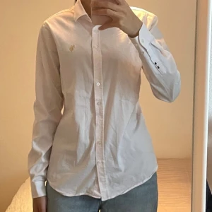 Vit skjorta från Ralph Lauren - Klassisk vit skjorta från Ralph Lauren med broderad logga i guld på bröstet. Skjortan har lång ärm, knappar framtill och en tidlös, normal passform. Perfekt till jeans eller kostymbyxor för en clean och stilren look.