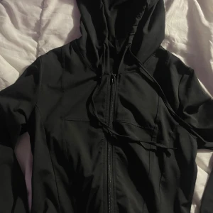 Svart hoodie med dragkedja i S - Svart hoodie med hel dragkedja framtill och snörning i huvan. Tillverkad i mjuk och stretchig polyesterblandning med 8% spandex för extra komfort. Perfekt för chill dagar eller när du vill ha en enkel och clean look.