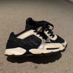 Chanel sneakers svart och vit - Äkta Snygga Chanel sneakers i svart, vitt och silver med klassisk CC-logga på sidan och Chanel-broderi bak. Skorna har snörning, rund tå och platt sula. Materialmix av skinn och textil ger en lyxig känsla och cool kontrast. Kommer med låda och dustbag