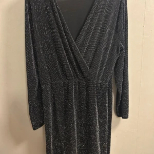 Svart glittrig långklänning omlott - Svart långklänning med glittriga detaljer och omlott-v-ringning. Klänningen har långa ärmar och ett lätt stretchigt material som ger en snygg passform. Perfekt för dig som vill sticka ut med en elegant och trendig look. Perfekt till nyår. One size men passar en L/Xl/XXL
