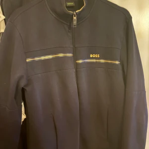 Svart ziptröja från BOSS med gula detaljer - Svart långärmad ziptröja från BOSS med gul logga på bröstet och tunna gula och blå linjer över bröstet. Tröjan har hög krage och hel dragkedja framtill. Perfekt för en stilren och sportig look.